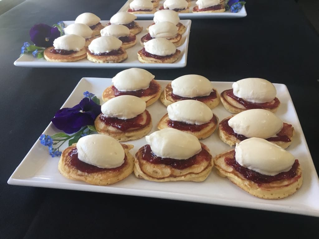 Two plates of nine pikelets with jam and cream on top, and some spring flowers decorating the square plates they're served on